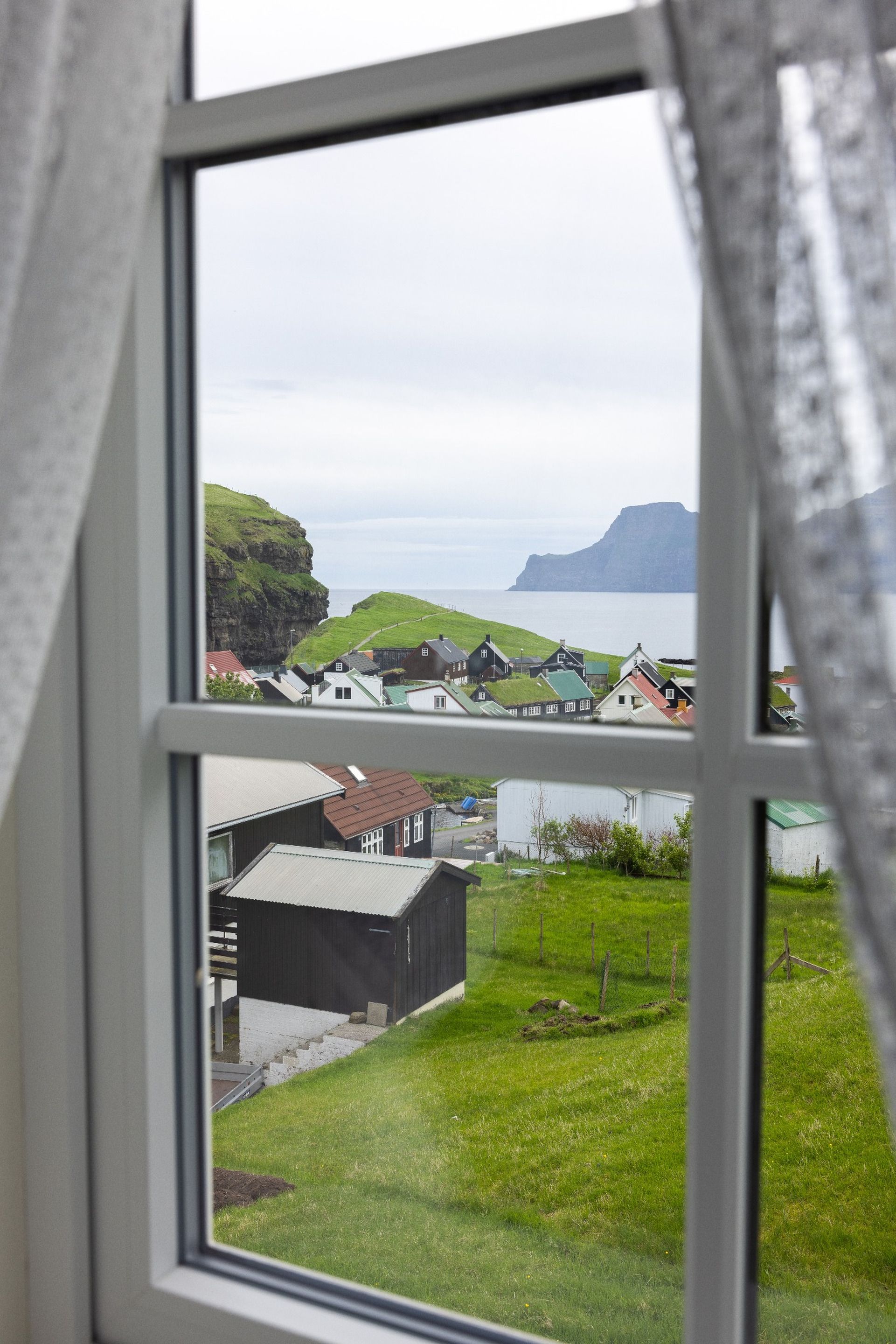 En utsikt från ett fönster över en kustby med hus, gräsbevuxna fält och en klippa.