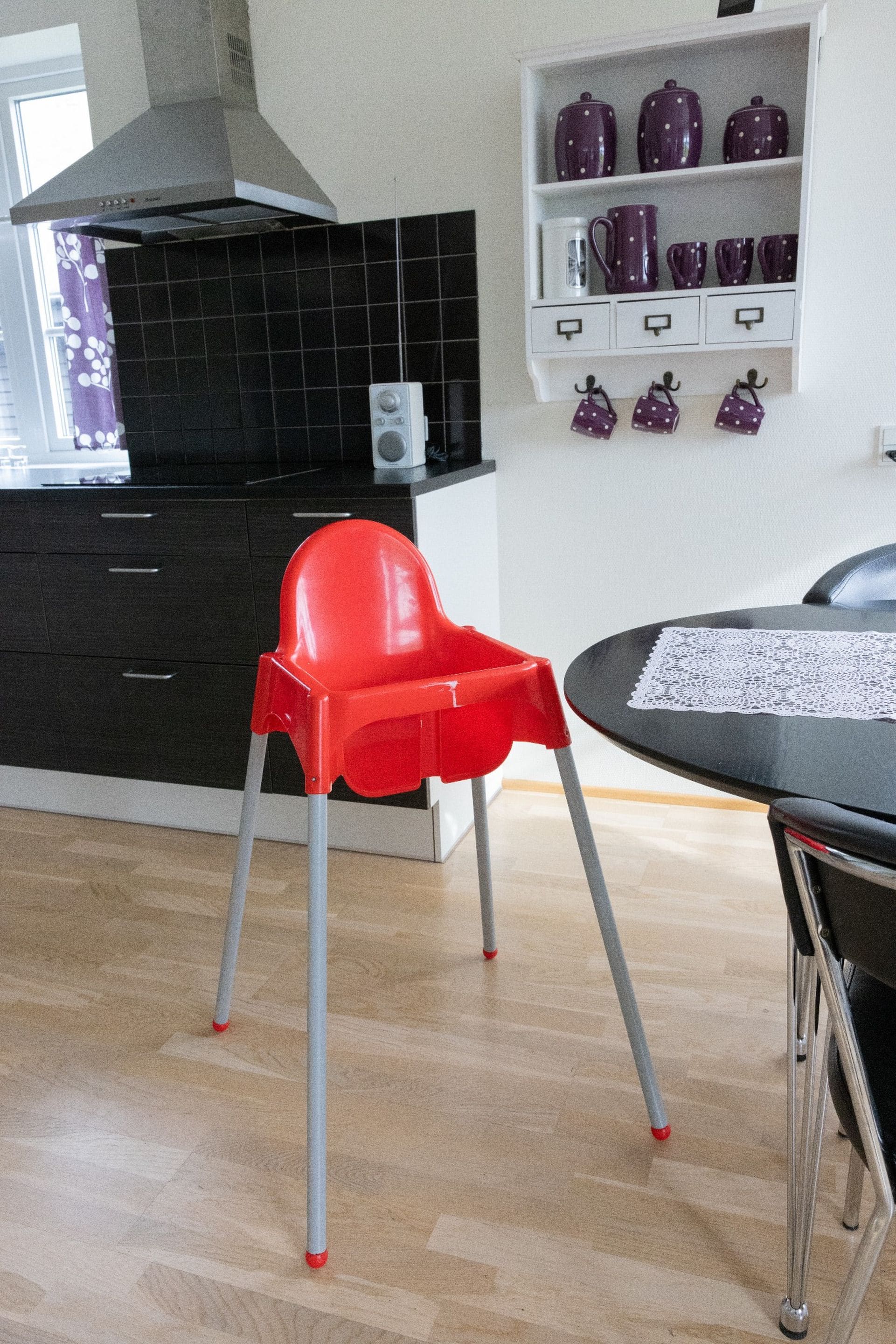 Una cocina moderna con una trona roja, armarios negros y un estante blanco con tazas moradas.