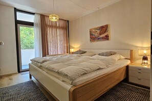 A bedroom with a bed, a nightstand, a lamp, a painting on the wall, and a view through a window. - Holiday apartment with 1 bedroom (Bad Harzburg)