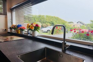 A kitchen with a stainless steel sink, a window with blinds, and a view of a car and greenery outside. - Holiday home with 2 bedrooms (Julianadorp)