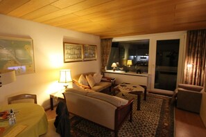 A cozy living room with a sofa, a coffee table, and a dining table set with a tablecloth. There is a window with curtains, a lamp, and a painting on the wall. - Holiday apartment with 1 bedroom (Cuxhaven)