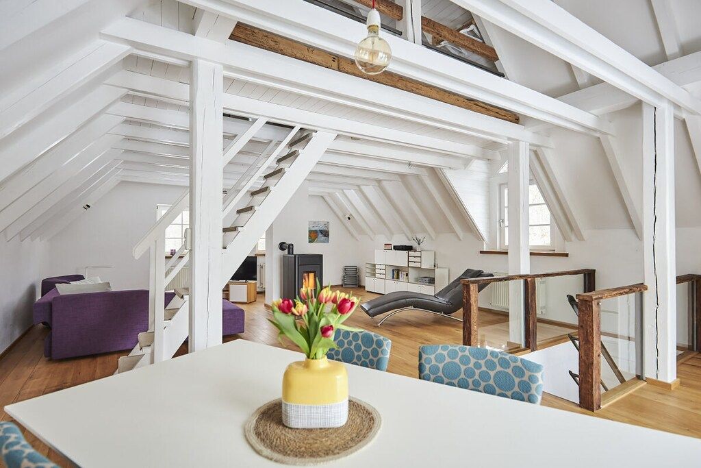 A spacious loft with a high ceiling, exposed wooden beams, a dining area with a white table, and a staircase leading to an upper level.