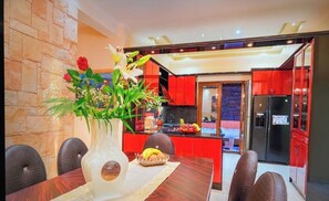 A modern kitchen with red cabinets, a black refrigerator, and a dining area with a vase of flowers and fruit. - Luxury Villa in Corfu with Indoor jaccuzi 15 min walk to town - Villa Foni Corfu (Kerkira)