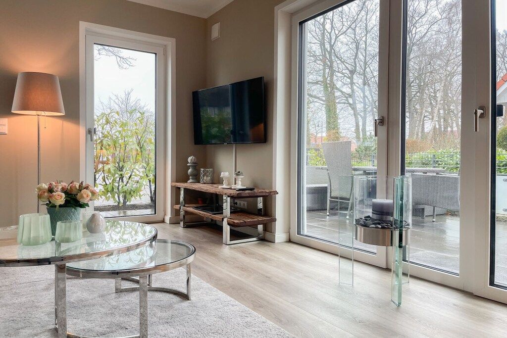 A modern living room with a glass coffee table, a flat-screen TV mounted on the wall, and a sliding glass door leading to an outdoor area.