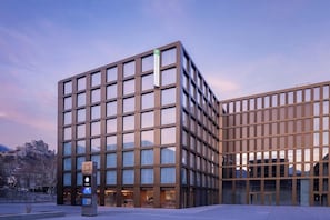 A modern building with a glass facade, a sign indicating a cafe, and a view of a mountain in the background. - Holiday Inn Express and Suites Sion By IHG (Sion)