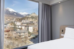 Una habitación de hotel con un amplio ventanal que ofrece vistas a un paisaje montañoso y un edificio histórico.