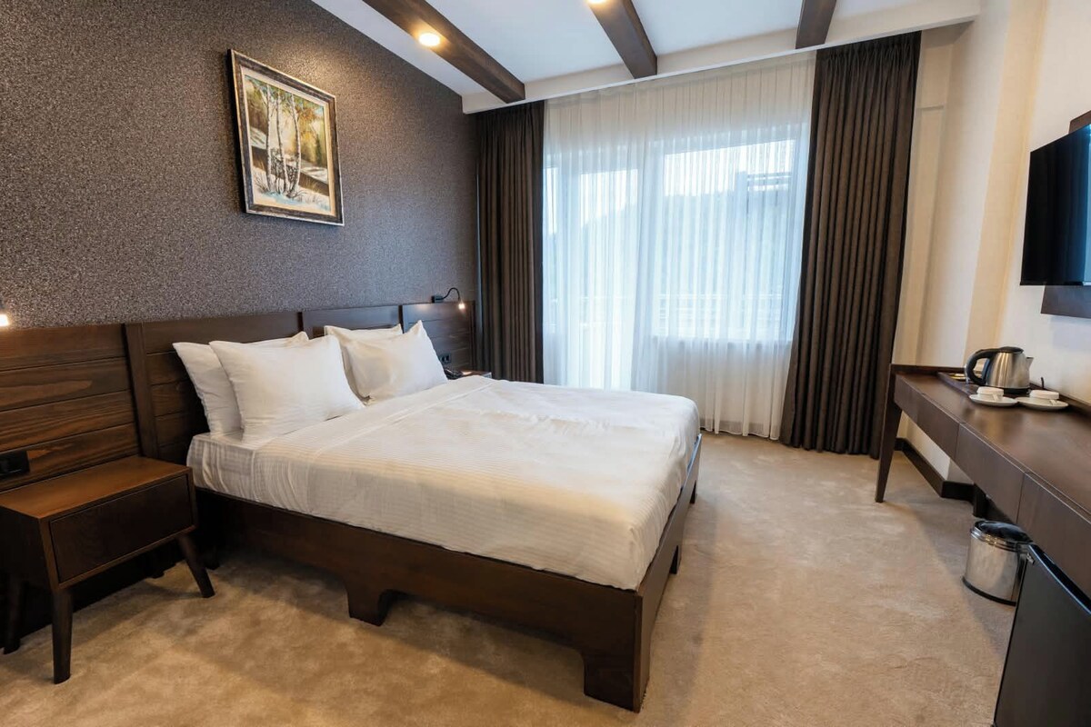 A hotel room with a large bed, a bedside table, a desk, a chair, a television, and a window with curtains.