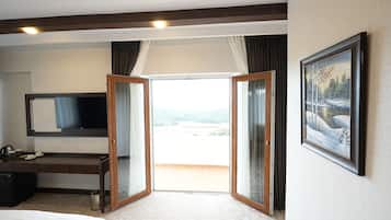 A hotel room with a bed, a desk with a flat-screen TV, a painting on the wall, and a balcony with a view.