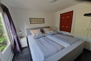 A double bed with striped bedding, a nightstand with a lamp, a framed picture on the wall, and a window with curtains. - Holiday apartment with 1 bedroom (Callantsoog)