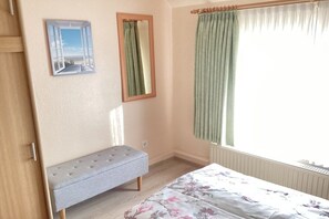 A bedroom with a bed, a bench, a window with curtains, and a mirror. - Holiday apartment with 1 bedroom (Cuxhaven)