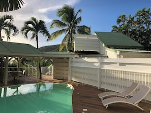 A pool area with lounge chairs, a covered seating area, and palm trees in the background. - Tropikal Cottage. Sea view in Deshaies. Swimming pool. (Deshaies)
