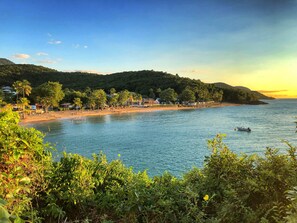 A coastal beach with a row of houses along the shore, a boat on the water, and dense greenery in the foreground. - Villa O Coeur de Deshaies. Sea view. Swimming pool (Deshaies)