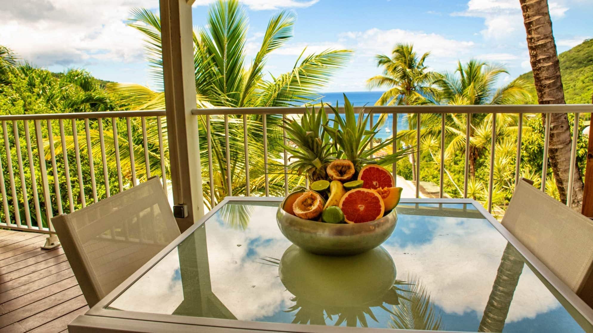 A glass-top table with a bowl of fruit on a balcony with a view of palm trees and the ocean.