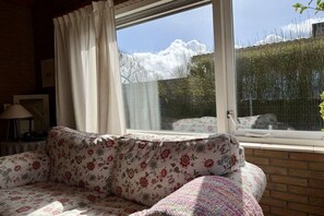 Ein Zimmer mit einer Couch, einem Fenster mit Vorhängen und Blick auf Bäume und einen Zaun.