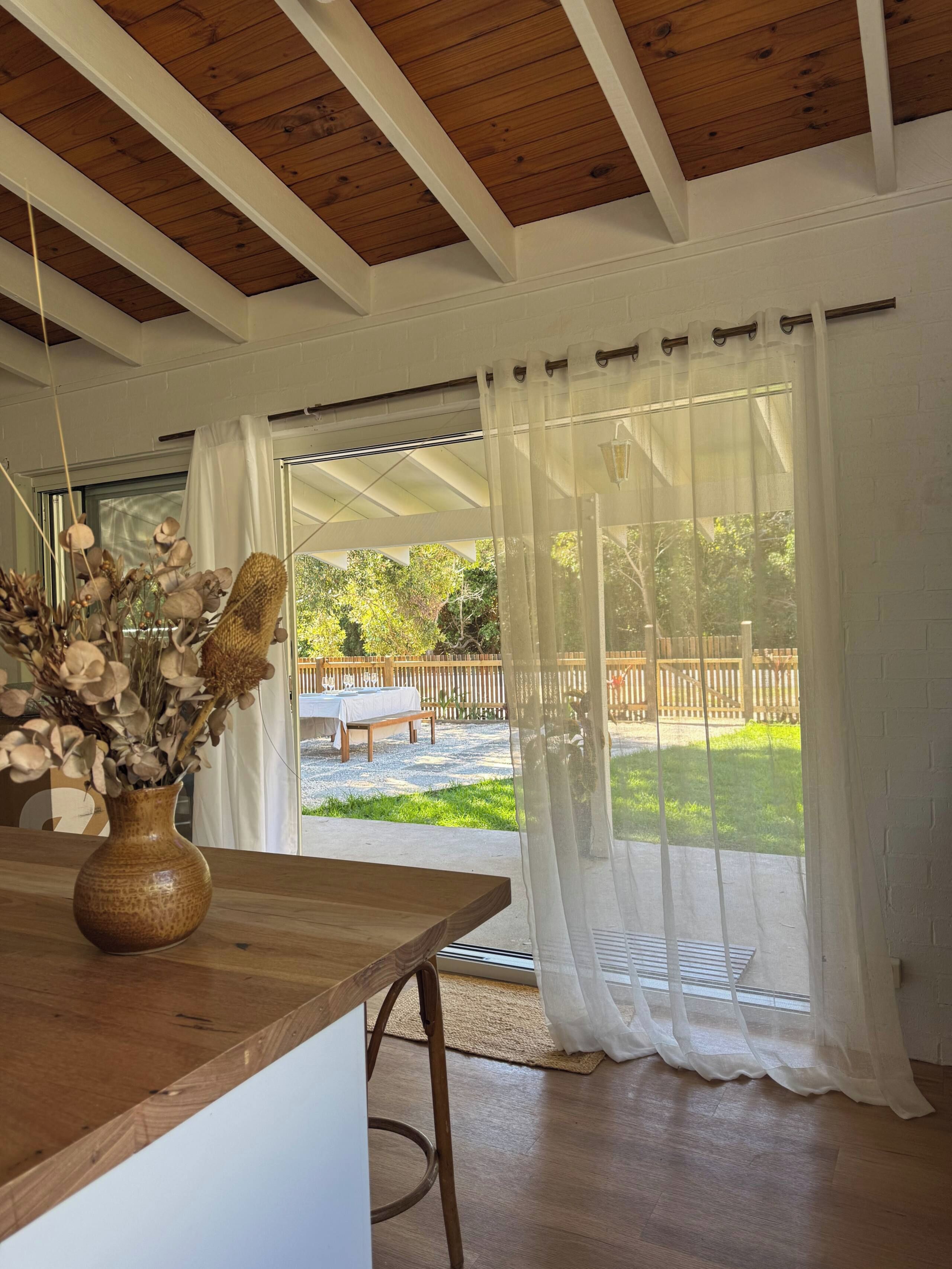Une pièce avec un plafond en bois, une table ornée d’un vase de fleurs séchées et une vue sur une terrasse extérieure à travers de fines rideaux.