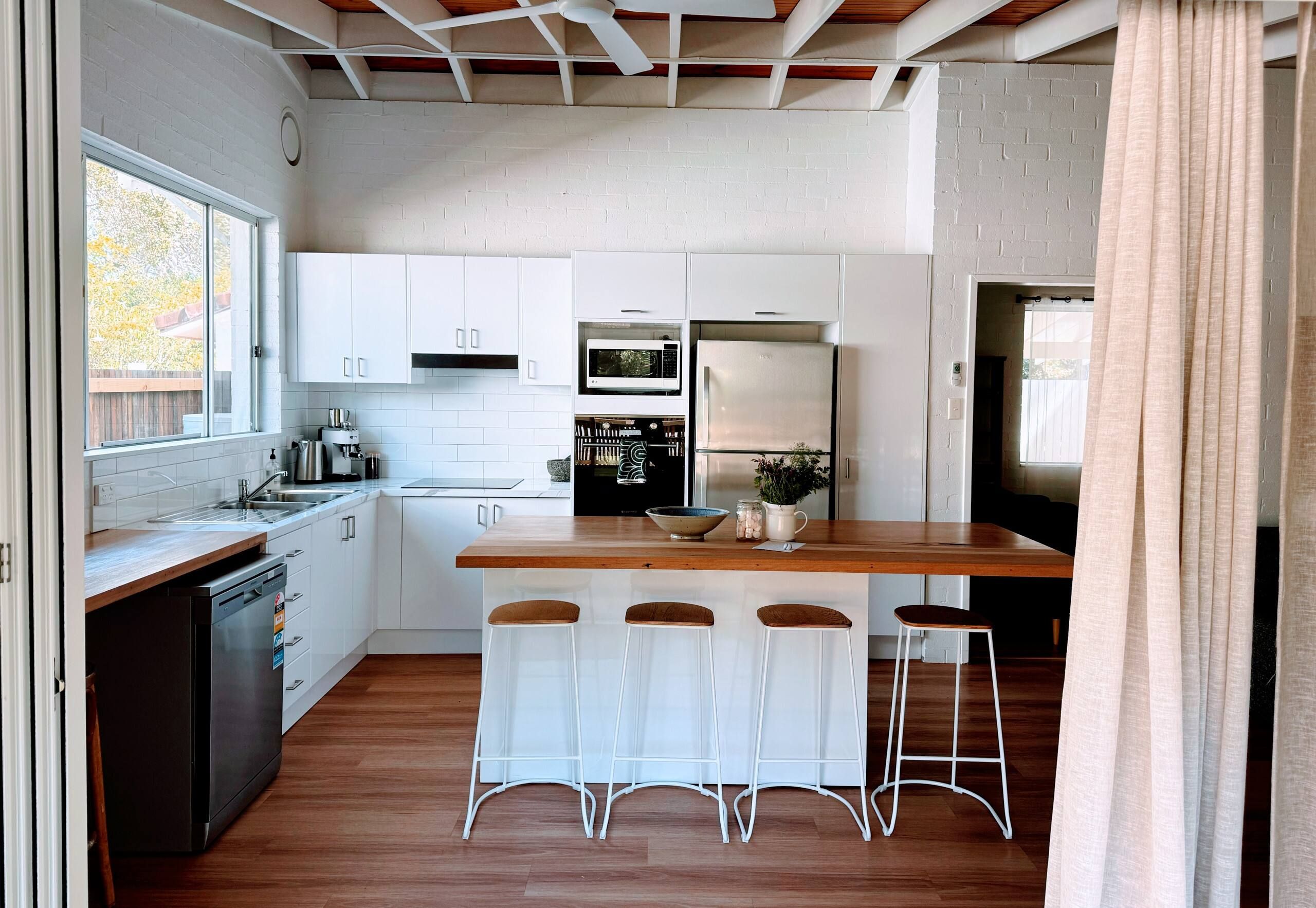 Une cuisine moderne avec des meubles blancs, un plan de travail en bois, un four à micro-ondes encastré et un réfrigérateur. On y trouve quatre tabourets de bar et une fenêtre qui laisse entrer la lumière naturelle.