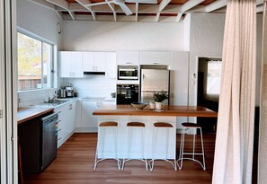 Cozinha moderna com armários brancos, bancada de madeira, micro-ondas embutido e geladeira. Há quatro banquetas e uma janela que oferece iluminação natural.