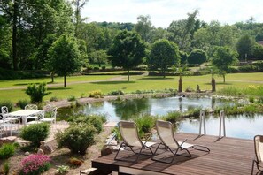A serene outdoor setting with a pond, deck chairs, and a table, surrounded by lush greenery and trees. - Castle in Buttlar with swimming pool (Buttlar)