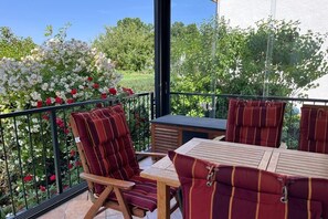 A balcony with a wooden table and two striped cushioned chairs, surrounded by a metal railing and a garden with white and red flowers. - Holiday apartment with 2 bedrooms (Eschwege)