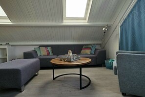 Ein Loft-Zimmer mit hoher Decke, Dachfenster, grauem Sofa mit bunten Kissen, einem runden Holztisch und einem grauen Sessel.