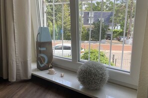 Een raam met uitzicht op een straat, een plankje met decoratieve spullen, en een gedeeltelijk open gordijn.