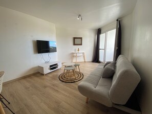 Un salon avec un canapé, un téléviseur fixé au mur, une petite table avec des chaises et une fenêtre avec des rideaux.