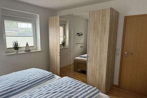 A bedroom with a bed, a wardrobe, a mirror, and a window with plants. - Holiday apartment with 1 bedroom (Borkum)