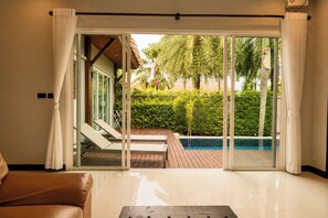 A room with a sliding glass door leading to a pool area, a wooden deck, and a view of a lush garden.