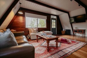 Ein gemütliches Schlafzimmer im Loft-Stil mit schrägem Dach, einem großen Fenster, Holzboden, einer Couch, einem Couchtisch und einem Fernseher.