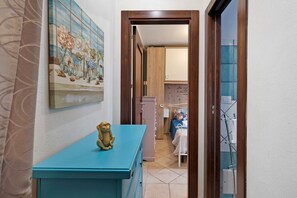 A hallway with a blue cabinet, a wall-mounted painting, and a wooden door leading to a room with a bed and a towel rack. - Apartment 'Cala Bianca – Giardino & Relax a Torre Lapillo' (Porto Cesareo)