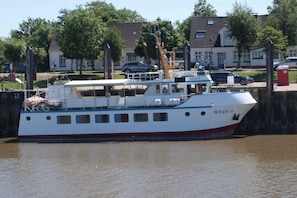 A white and red boat docked at a pier with a house and cars in the background. - Holiday home with 2 bedrooms (Butjadingen-Stollhamm)