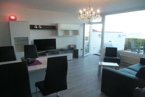 Een moderne woonkamer met een flatscreen televisie, een eethoek met stoelen, een kroonluchter en uitzicht door een glazen deur.