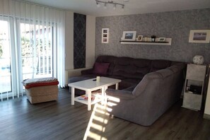 A living room with a grey sofa, a white coffee table, and a beige ottoman. There is a window with blinds, a wall-mounted shelf with pictures, and a small cabinet. - Holiday home with 1 bedroom (Wurster Nordseeküste)