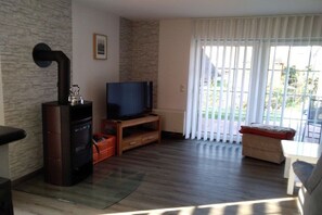 A living room with a fireplace, a television on a wooden stand, and a large window with blinds. - Holiday home with 1 bedroom (Wurster Nordseeküste)