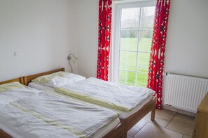Quarto com duas camas de solteiro, janela com cortinas vermelhas, radiador e vista para um gramado verde.