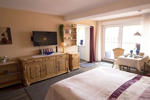 A hotel room with a bed, a television, a wooden cabinet, a small table with a chair, and a window with curtains. - - Heidi - Apartment Sea Sounds (Borkum)