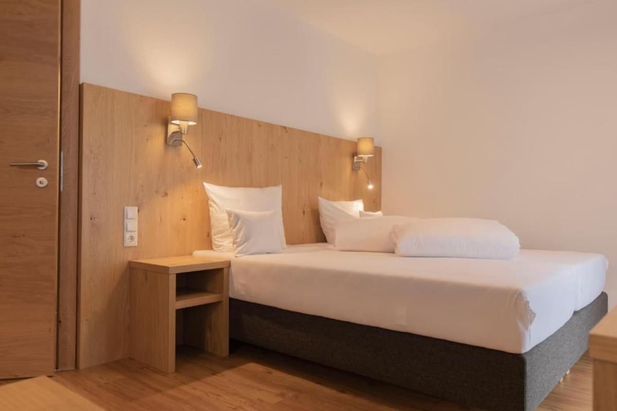 Une chambre d’hôtel avec une tête de lit en bois, deux lampes de chevet, un lit avec du linge de maison blanc et une petite table de chevet en bois.