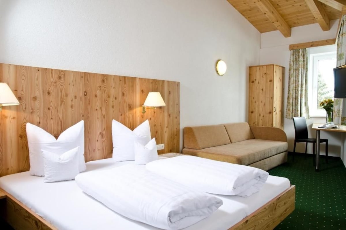 Une chambre d’hôtel avec une tête de lit en bois, deux lits, un canapé, un bureau avec une chaise et une fenêtre avec des rideaux.