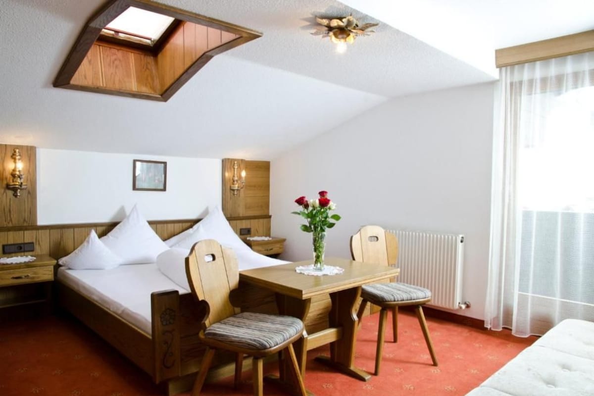 Une chambre d’hôtel avec un lit, une table en bois et deux chaises, un vase avec des fleurs et une lucarne.