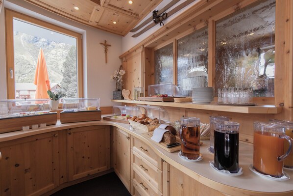 A wooden interior with a breakfast bar, a cross on the wall, and a window with a view of a mountain. - Berghotel Rasis (Galtür)