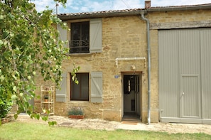 Un bâtiment rustique en pierre, doté d’un grand portail de garage, d’une enseigne portant le numéro « 13 » et d’un petit jardin agrémenté de plantes en pot.