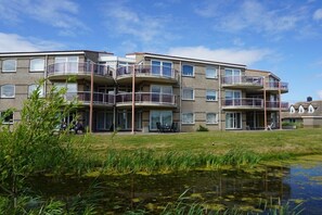 A multi-story residential building with balconies, a grassy area, and a water feature in the foreground. - Residence Juliana 19 (Julianadorp)