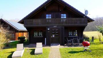 Een houten chalet met twee verdiepingen, een balkon, nummer 207 en een barbecue in de tuin.
