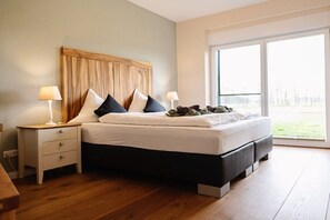 A bedroom with a large bed, two bedside tables with lamps, a wooden headboard, and a view through a large window.