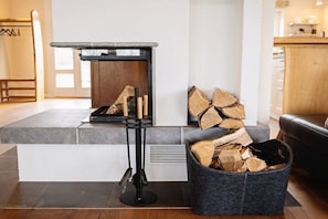 A modern living room with a fireplace, a black stand with logs, and a grey felt bin with firewood.