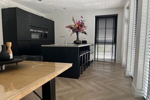 A modern kitchen with black cabinetry, a wooden dining table, and a large window with blinds. - Villa in Julianadorp with barbecue (Julianadorp)