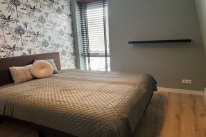 A bedroom with a bed, a window with blinds, a wall-mounted shelf, and a patterned wallpaper. - Villa in Julianadorp with barbecue (Julianadorp)
