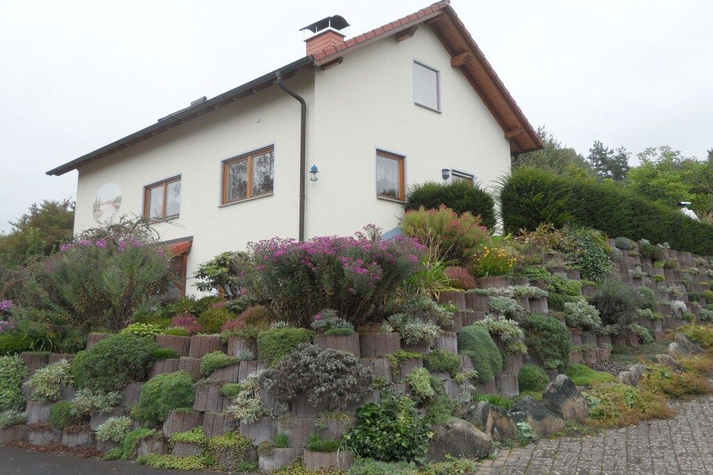 Une maison à deux étages, dotée d’un toit en pente, d’une cheminée en briques et d’un mur de soutènement en pierre agrémenté de diverses plantes.