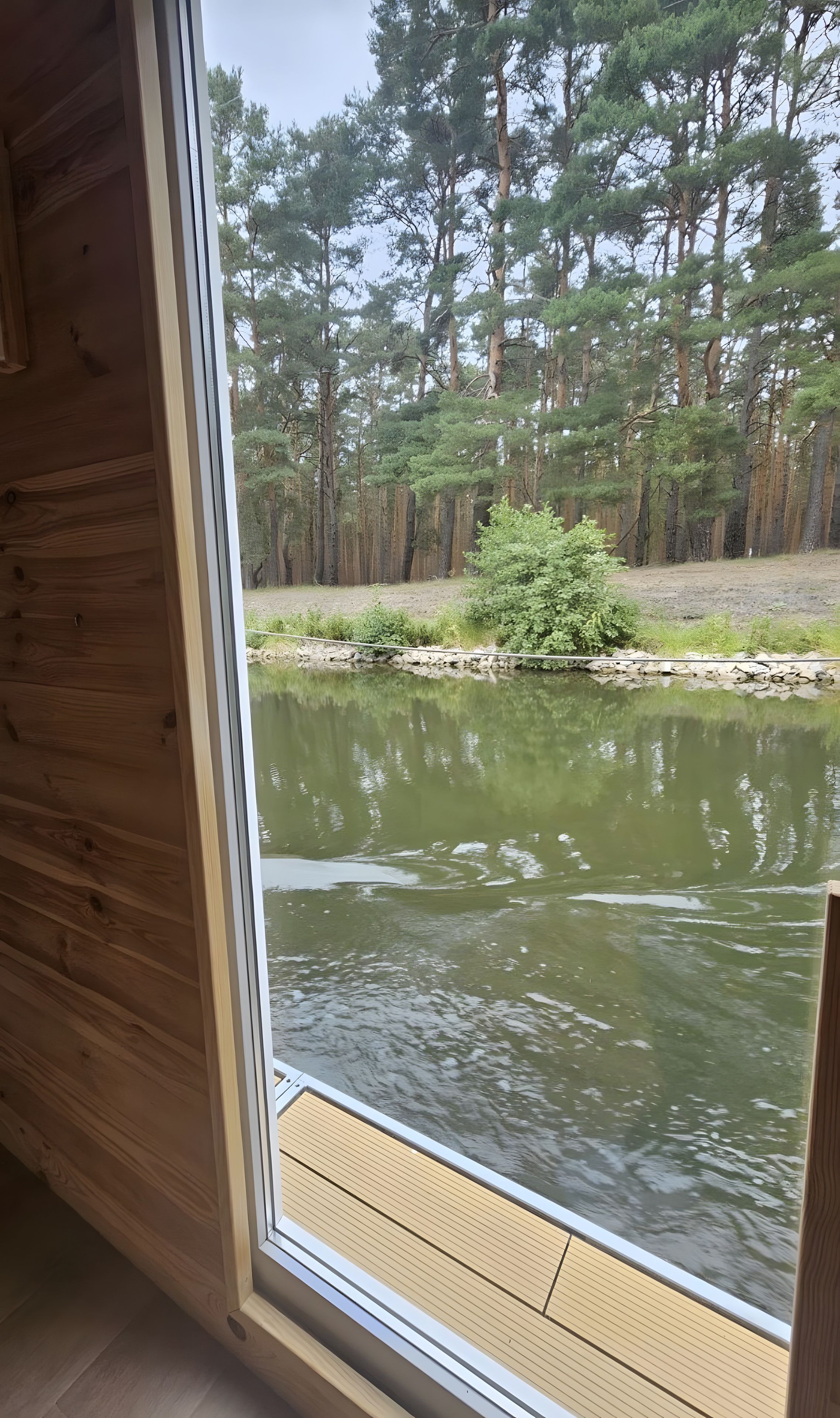 Blick aus einer Hütte mit Holzboden und Fenster auf einen Teich, der von Bäumen umgeben ist.