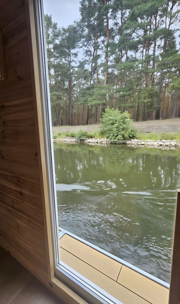 A view from inside a cabin with a wooden floor and a window overlooking a pond surrounded by trees.